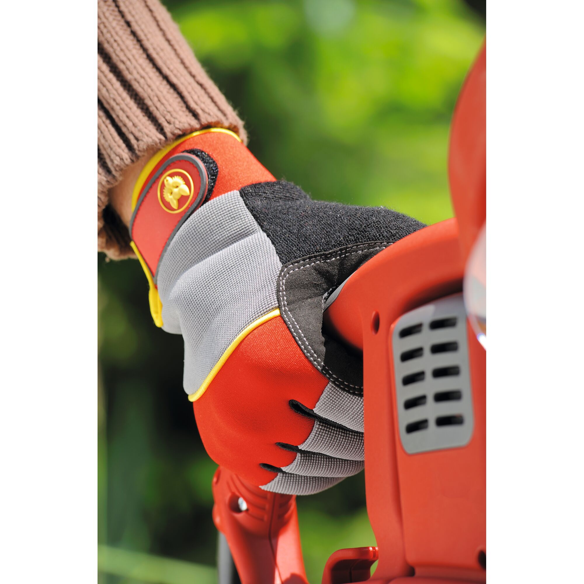 A close-up of a person wearing a red and grey WOLFGarten gardening glove, holding the handle of an orange garden tool with a blurred green background.
