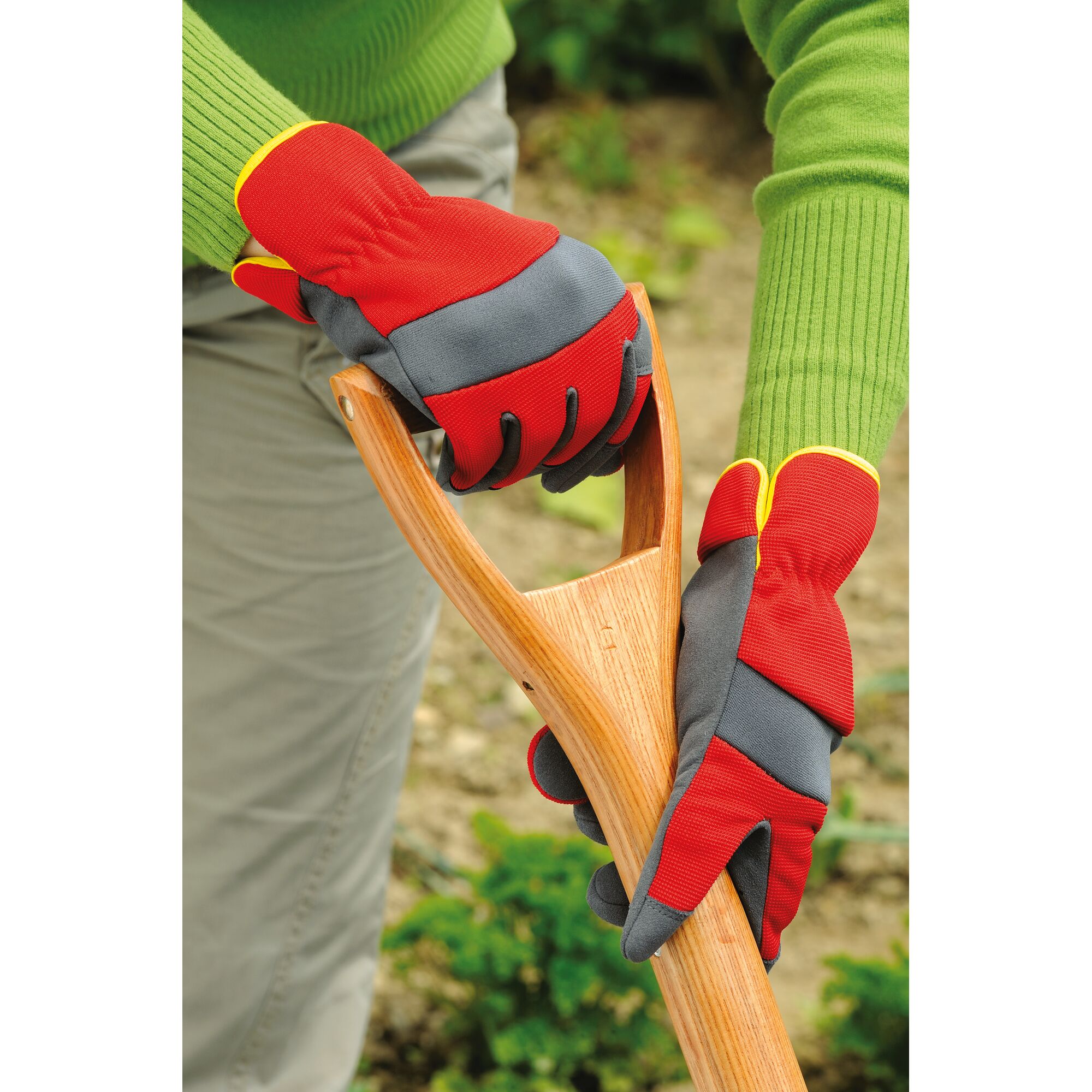 A person wearing red and grey gardening gloves is holding the wooden handle of a garden tool, possibly a spade or shovel, with a green sweater visible and a garden background.