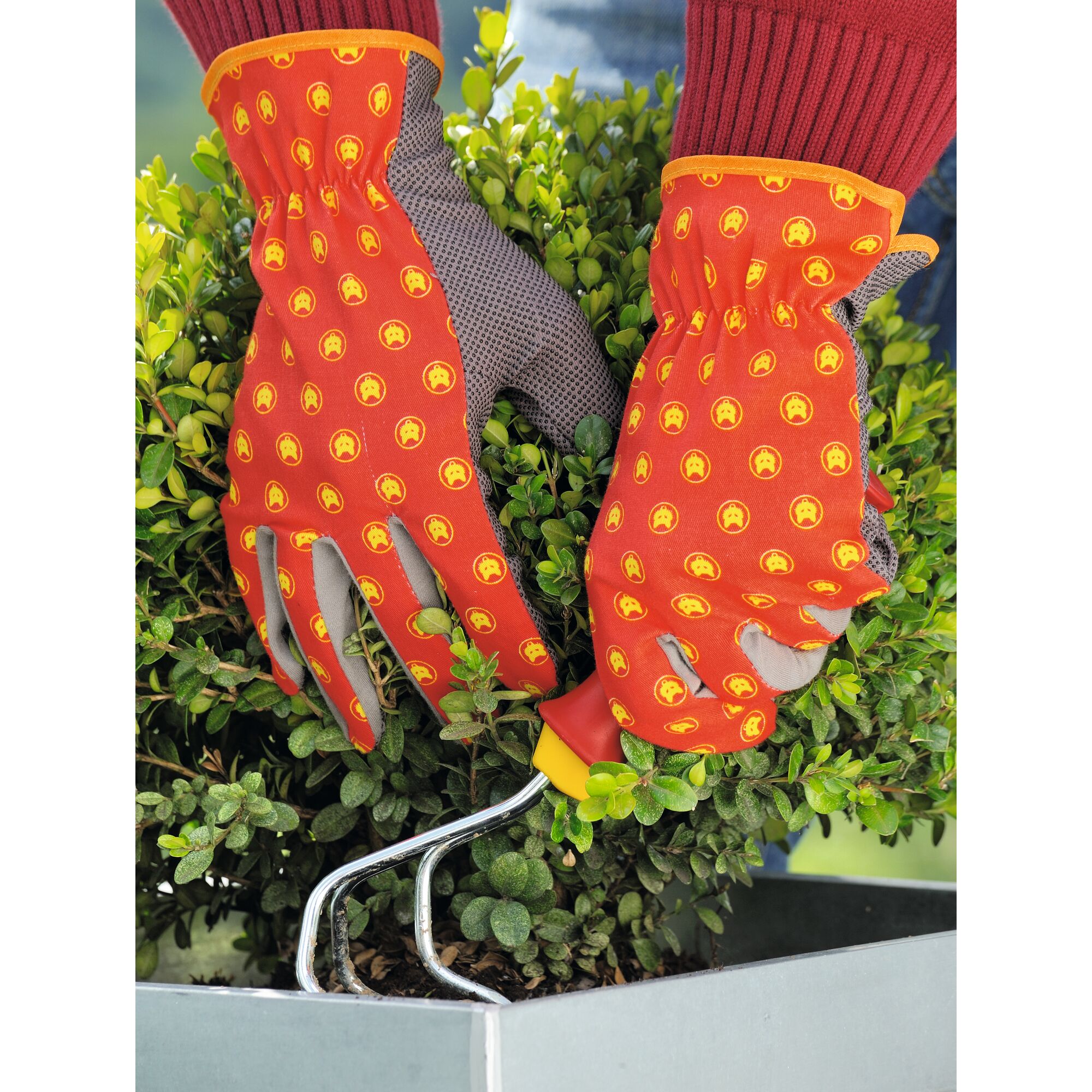 A person wearing orange-and-yellow patterned gardening gloves is using a small hand cultivator to work in the soil of a potted green shrub.