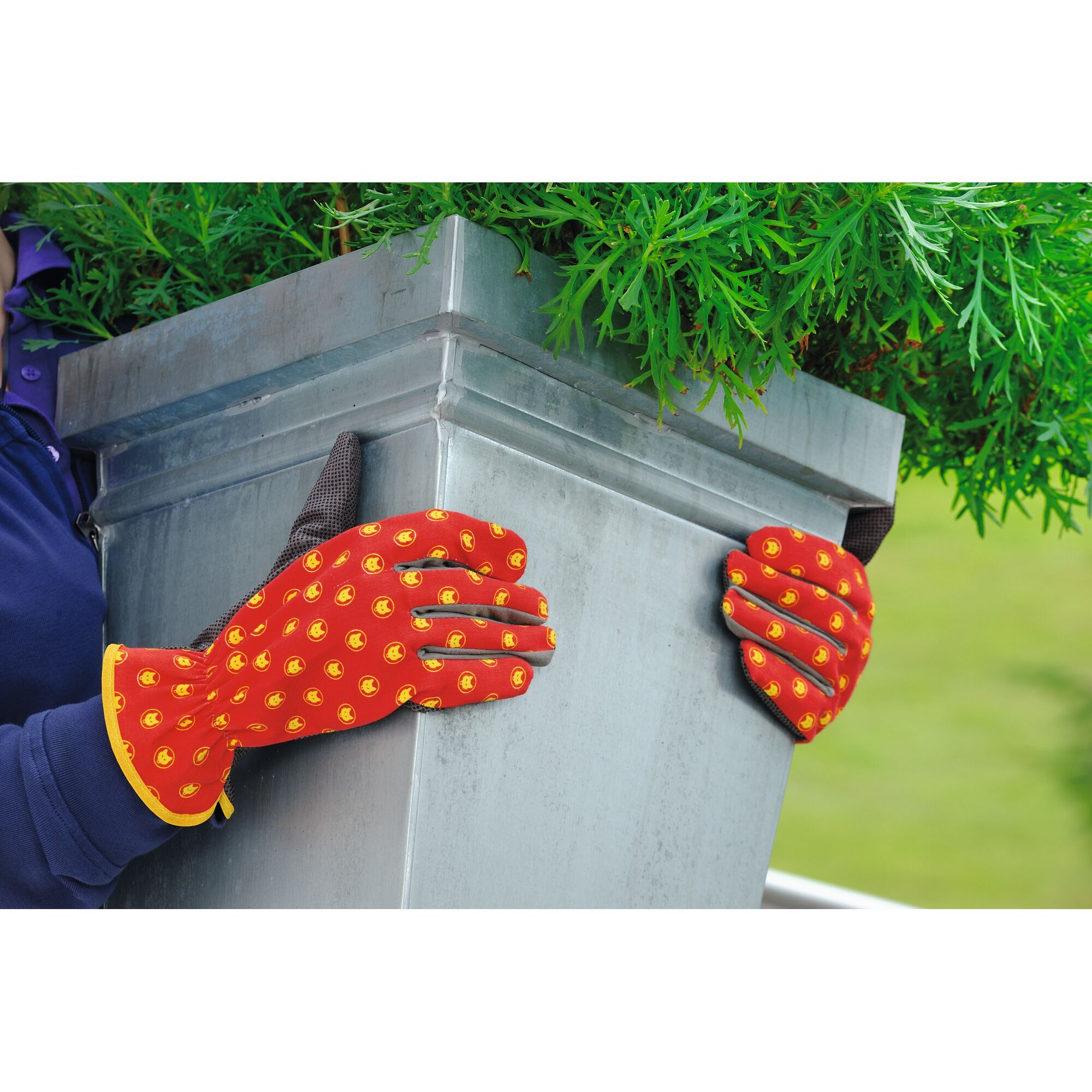 A person wearing red gardening gloves with yellow patterns is holding a large metal planter with green foliage.