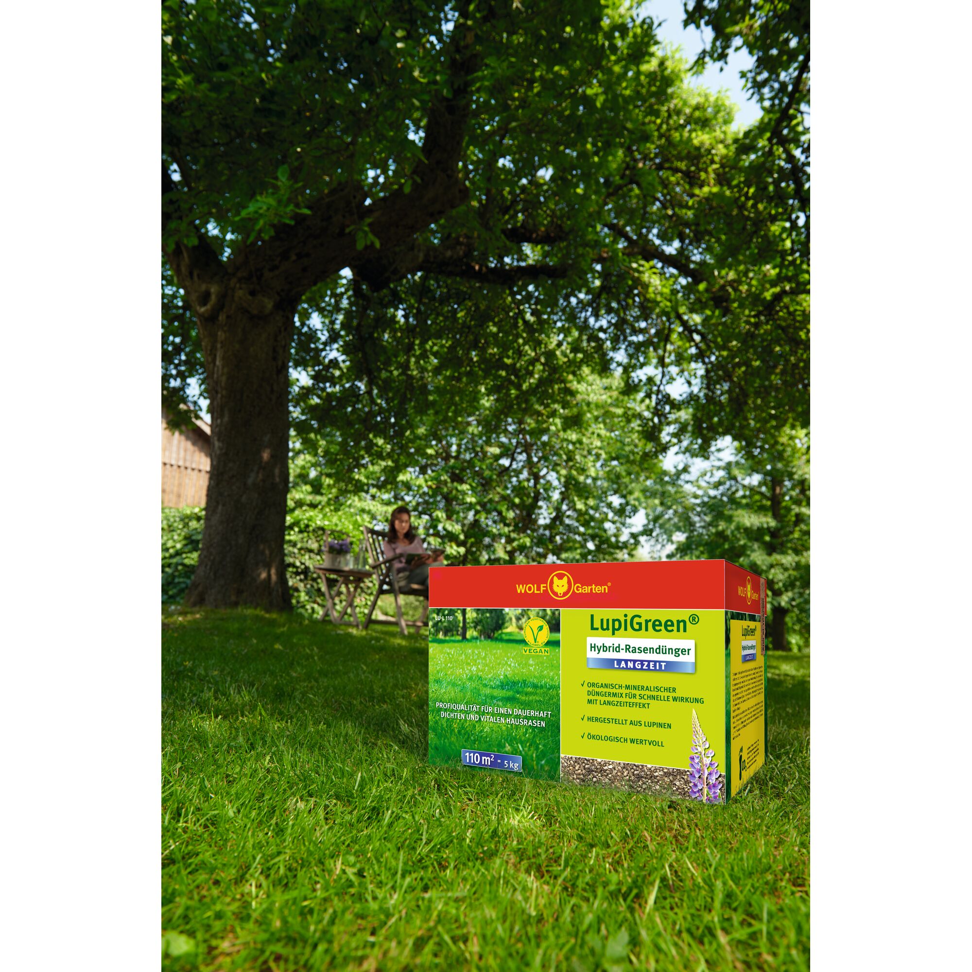 A box of WOLF-Garten LupiGreen hybrid lawn fertilizer is placed on a lush green lawn, with a large leafy tree and a person sitting at a garden table in the background.