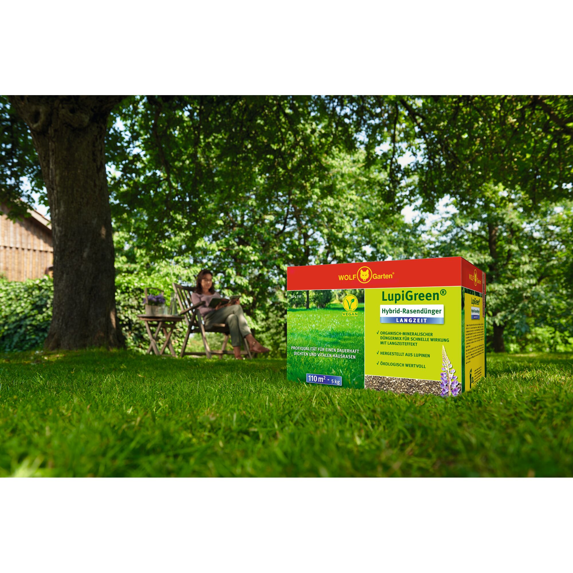 A box of WOLFGarten LupiGreen Hybrid Lawn Fertilizer is placed on a green lawn under a large tree in a garden. In the background, a person is sitting on a wooden chair reading, with their face blurred for privacy. The atmosphere is sunny and peaceful.