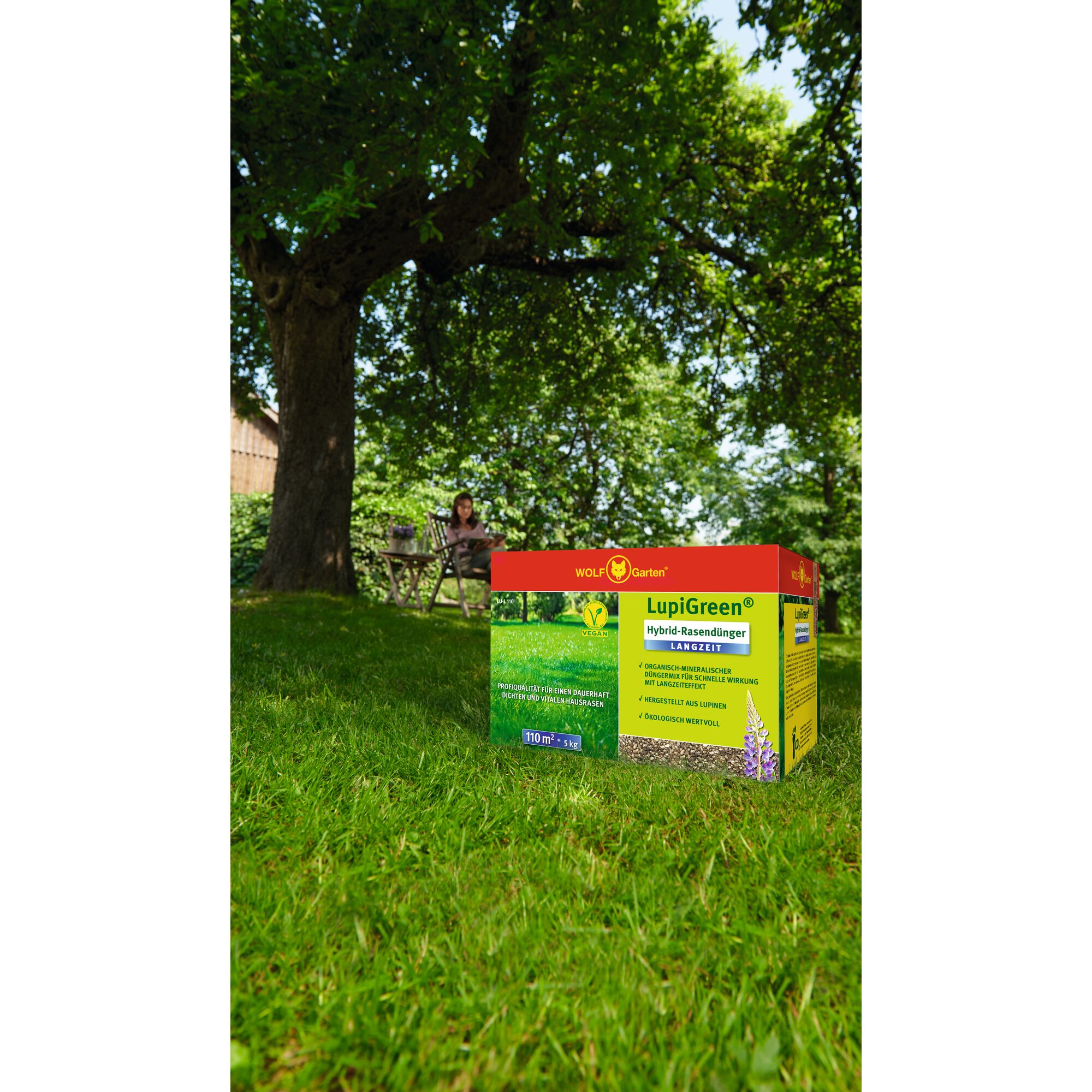 A box of WOLF Garten LupiGreen hybrid lawn fertilizer is placed on green grass in a garden with a large tree and a person sitting at a table in the background.