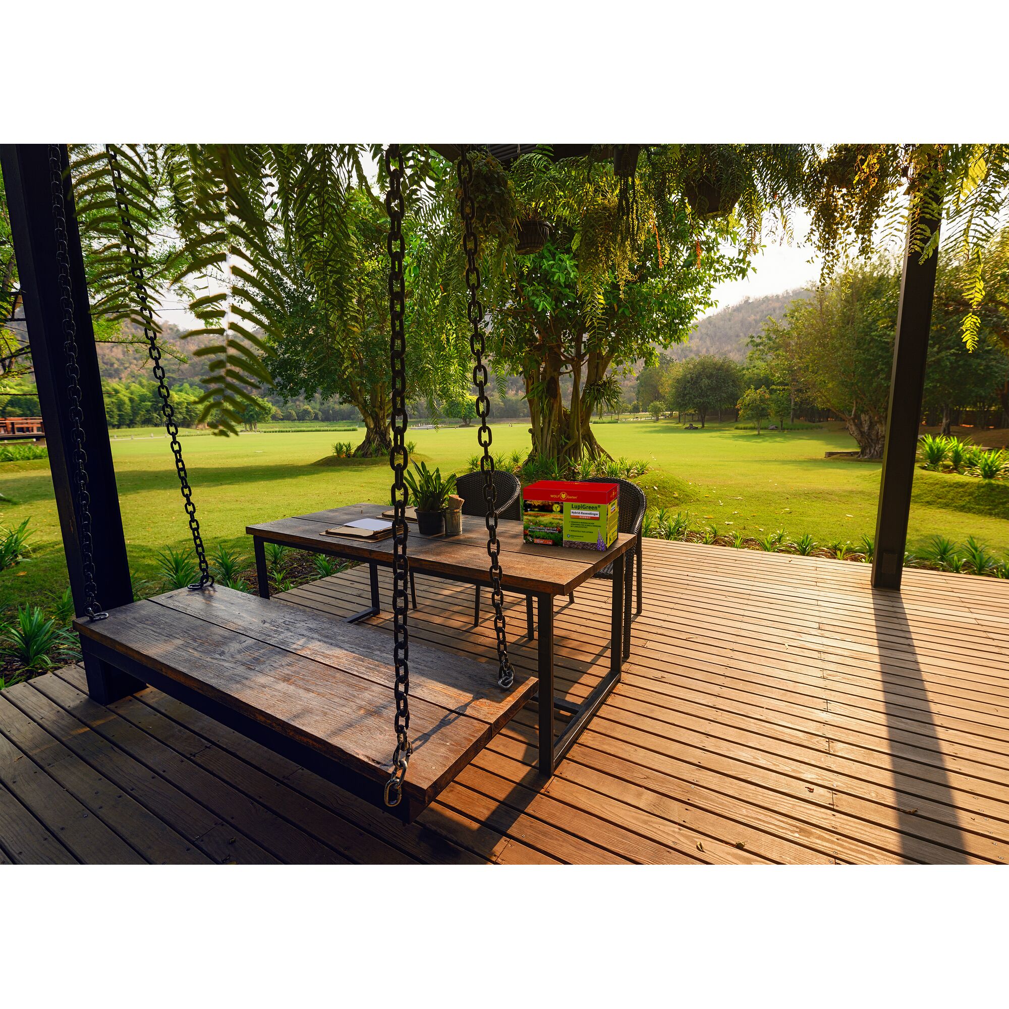 Wooden patio deck with a swing, a table holding gardening supplies including a branded WOLFGarten box, a notebook, and potted plants, overlooking a green garden with trees and sunlight.